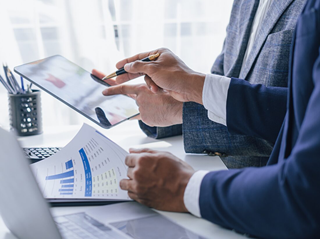 スーツを着たビジネスマンがタブレットを使ってデータを確認しながらディスカッションしている場面。