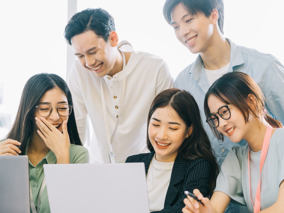 明るい室内で、5人の若いアジア人が笑顔でラップトップを囲んでいる。3人の女性が座り、2人の男性が立って覗き込んでいる。